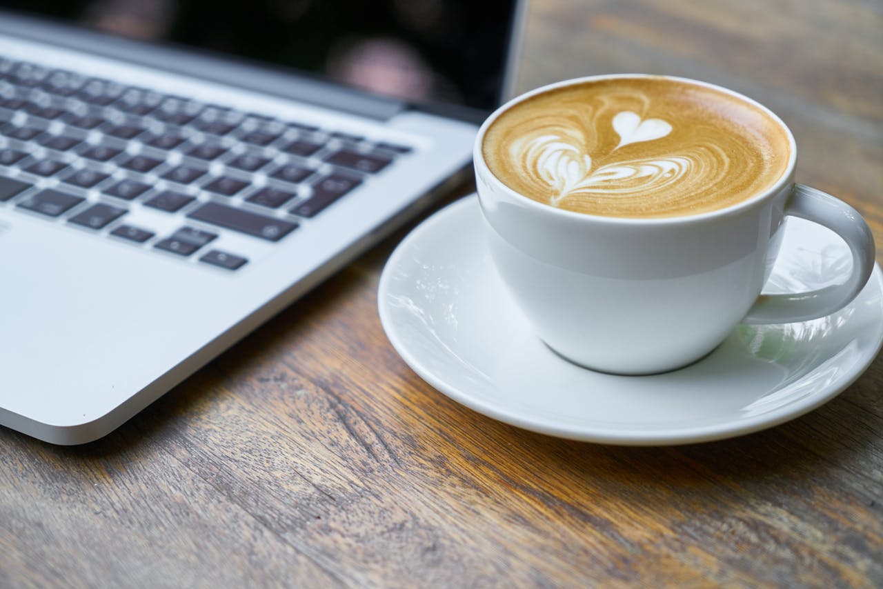 services-01 A cup of latte with intricate art beside a laptop on a wooden table, depicting a cozy work setting.